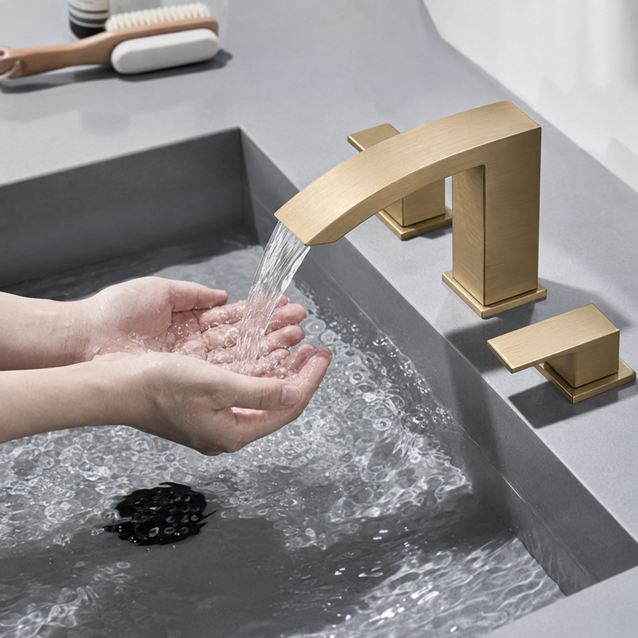 A modern matte gold faucet with a curved spout and matching side handles installed in a gray sink, with water running into a person's hands.