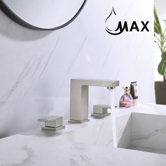 A modern matte silver two-handle bathroom faucet with running water, installed on a white marble sink against a white marble wall. A circular mirror is partially visible at the top, and a plant appears on the right.