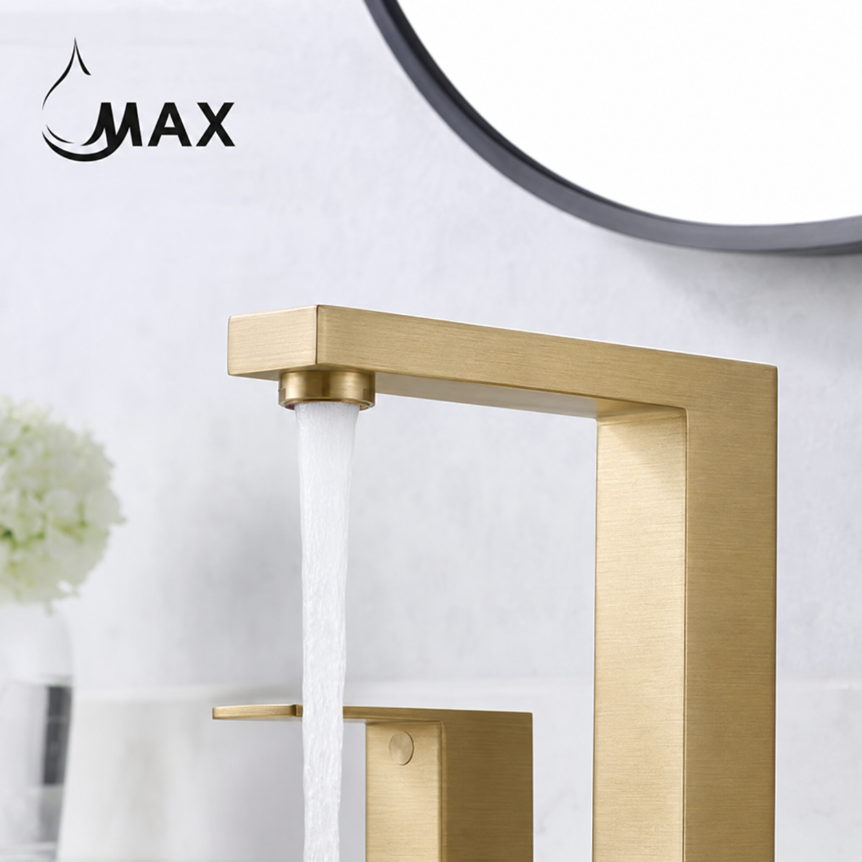 A modern rectangular faucet in brushed gold finish with water flowing from the spout, shown against a light bathroom wall. A circular mirror is partially visible at the top, and a small white floral arrangement appears at the left.