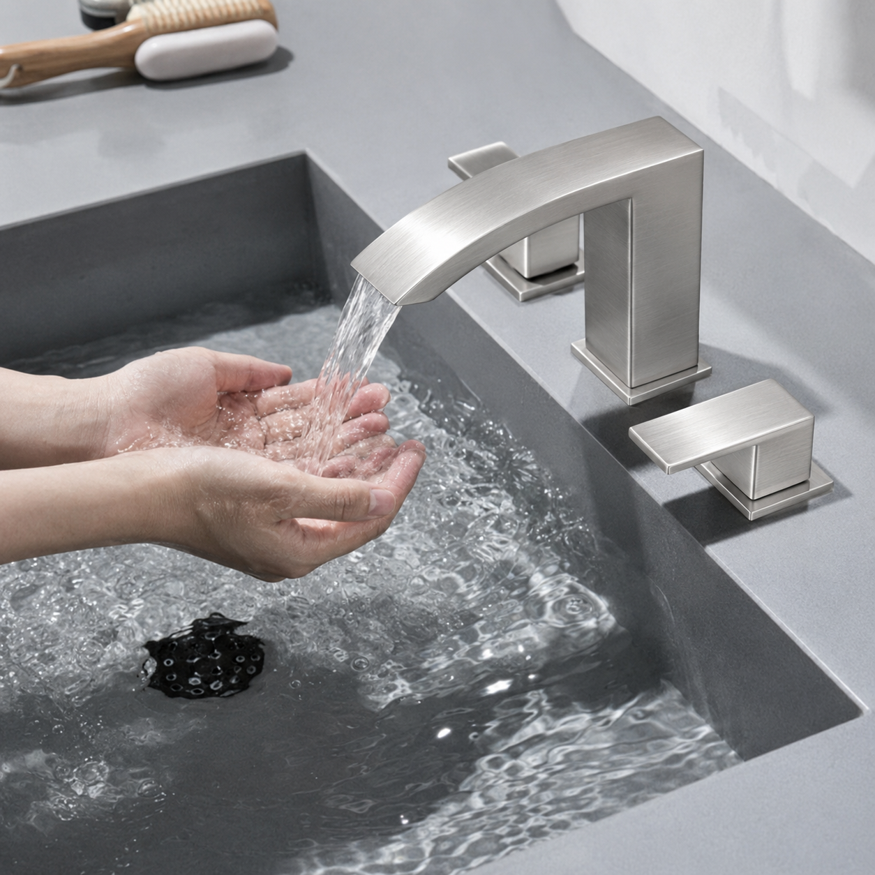 A modern brushed nickel faucet with a curved spout and two matching lever handles installed on a gray sink, with water running into a person's hands.