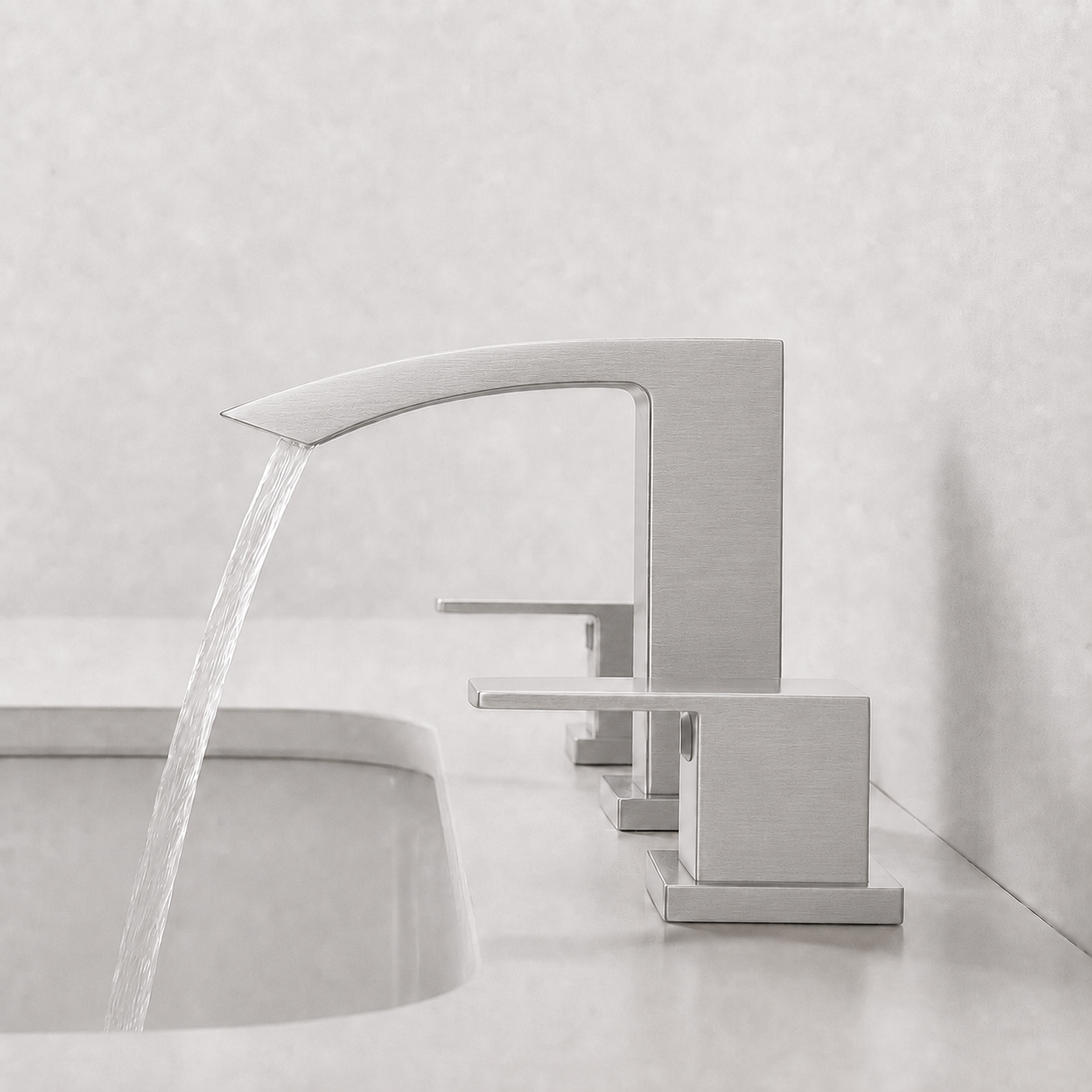 A modern matte silver waterfall bathroom faucet installed on a light countertop, with water flowing into a sink against a textured light gray wall.
