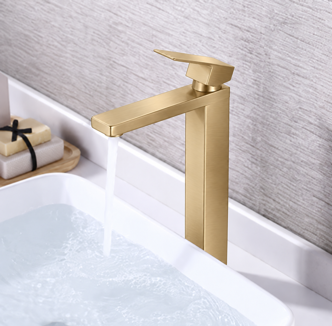 A modern single-handle tall bathroom faucet in brushed gold finish, shown with water running into a white sink against a textured gray wall.
